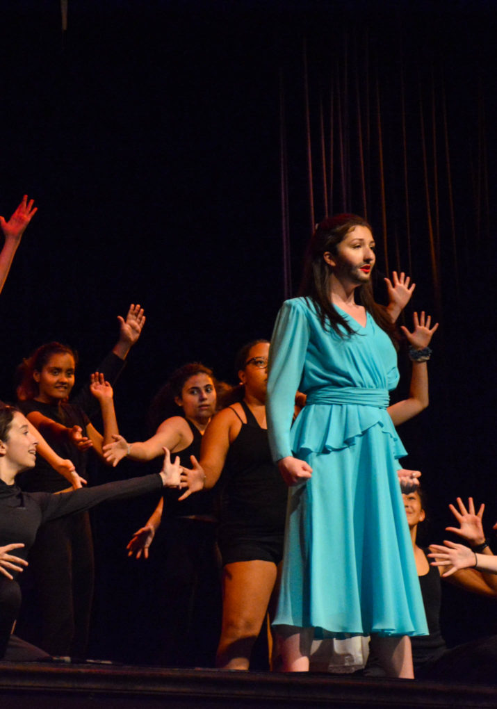 Camper in a blue dress with ensemble cast behind her on stage